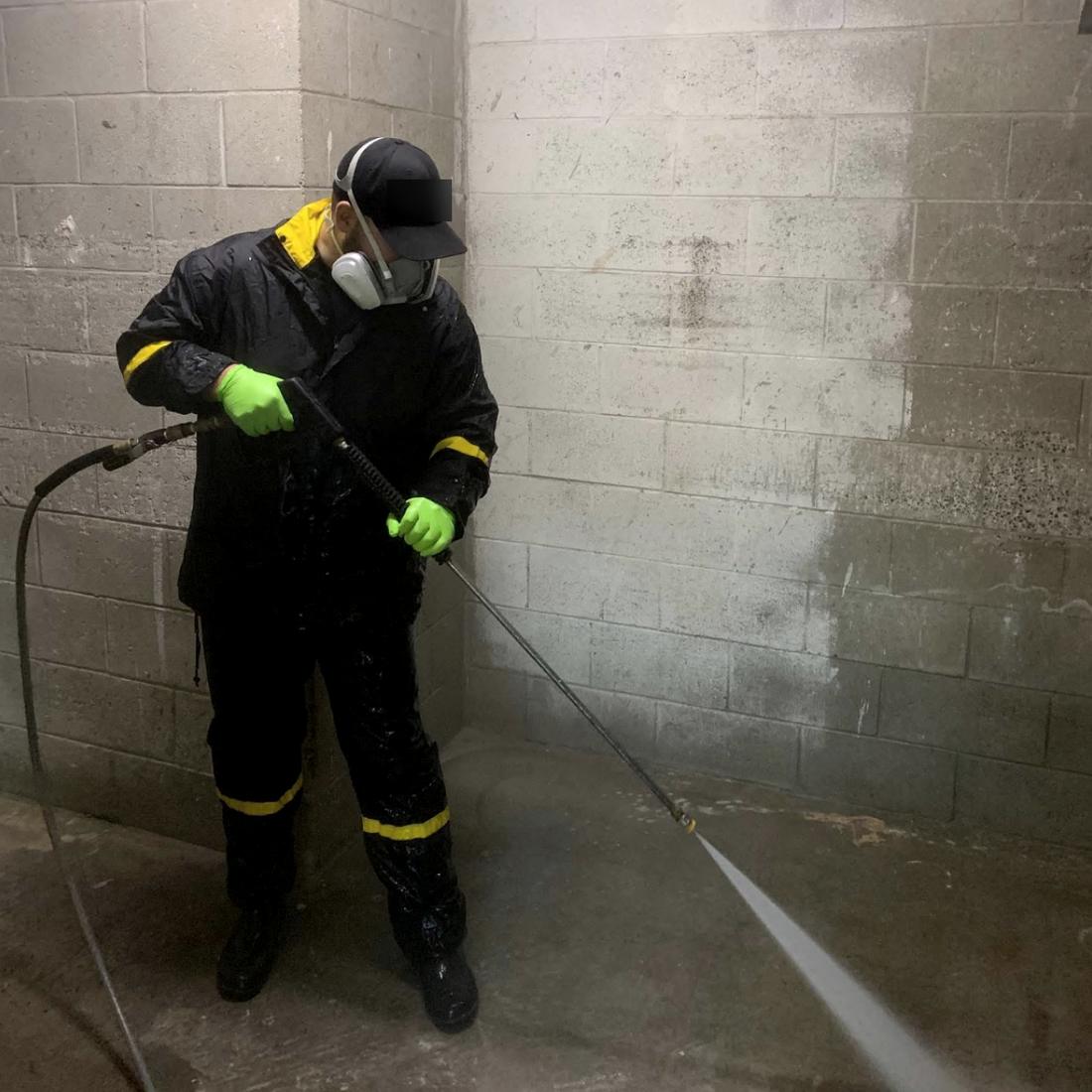 Technician in rain gear, respirator and gloves pressure washing a block wall