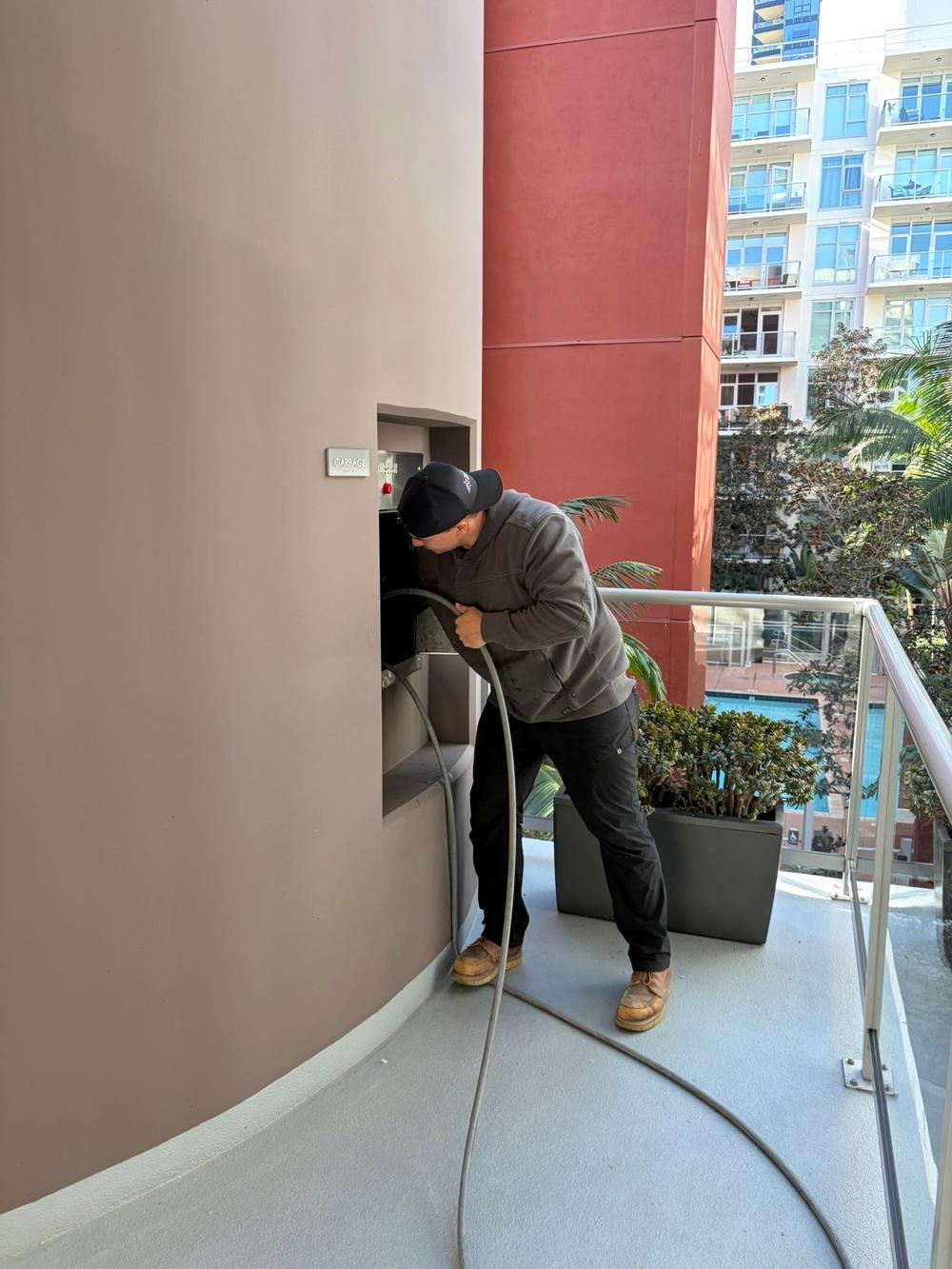 Technician feeding a pressure hose into a trash chute door on a high-rise terrace